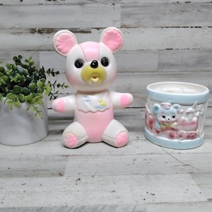 May include: A pink and white bear-shaped figurine with large black eyes and a yellow mouth. A light blue and pink ceramic planter with a bear and train design is next to the bear. A small white pot with green plants is also in the image.