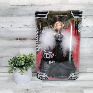 Puede incluir: Una muñeca Barbie rubia con un vestido negro con detalles de plumas blancas, exhibida en una caja de edición de coleccionista. La caja presenta un diseño de alfombra roja y el texto "Steppin' Out Barbie."