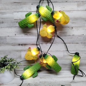 May include: A string of cactus-shaped fairy lights. The lights are green with yellow illumination, and have small black dots. The string is black, and the lights are hanging against a light-coloured wooden background.
