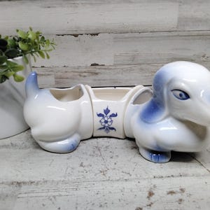 May include: A white ceramic dachshund-shaped condiment server with blue painted accents on the ears, tail, and eyes. The centre section features a blue floral pattern. The piece is divided into three distinct open compartments for serving.