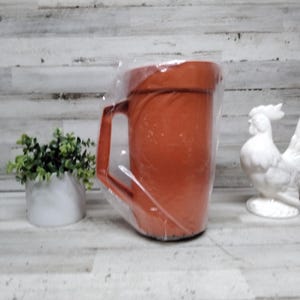 Può includere: Una grande brocca in ceramica arancione-rossa con manico, avvolta in plastica trasparente. Una statuetta di gallo in ceramica bianca e un piccolo vaso bianco con piante verdi sono rispettivamente a destra e a sinistra della brocca.
