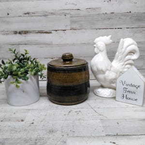 May include: A small green plant in a white pot, a brown and black ceramic jar with a lid, a white ceramic rooster figurine, and a white house-shaped sign with the text Vintage Tenn House printed in black script on the front.