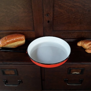 Könnte beinhalten: Eine rot-weiß emaillierte Pfanne mit schwarzem Griff liegt auf einer dunklen Holzoberfläche, neben einem Brotlaib und einem Croissant. Die Pfanne ist rund und leer. Das Brot ist goldbraun.