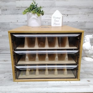 Puede incluir: Un mueble de almacenamiento de madera marrón claro con tres cajones horizontales con frentes de plástico transparente. Cada cajón contiene un inserto de plástico beige con múltiples divisores verticales para organizar objetos pequeños.