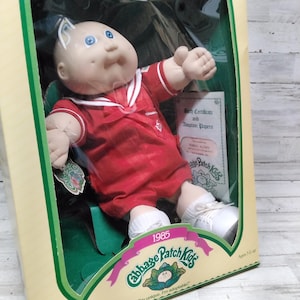 Puede incluir: Muñeca Cabbage Patch Kids vintage en su caja original. La muñeca viste un traje de marinero rojo con detalles blancos y zapatos. La caja muestra el logotipo de Cabbage Patch Kids y el año 1985.