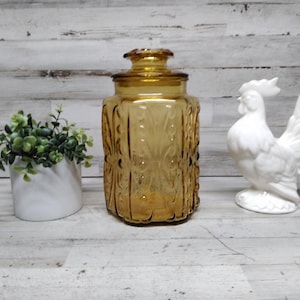 Op de afbeelding: Een amberkleurige glazen pot met een deksel, een witte keramische haanfiguur en een kleine witte plantenbak met groen. De pot heeft een decoratief reliëfpatroon. De haan staat rechtop.