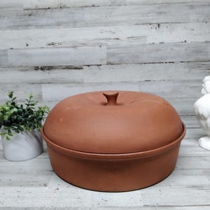 May include: A round, terracotta-colored ceramic baker with a lid and a small handle. The baker is designed for cooking and baking. The image also includes a white ceramic rooster figurine and a small potted plant.