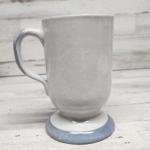 May include: A white ceramic mug with a blue band around the base. The mug has a handle and a rounded base.