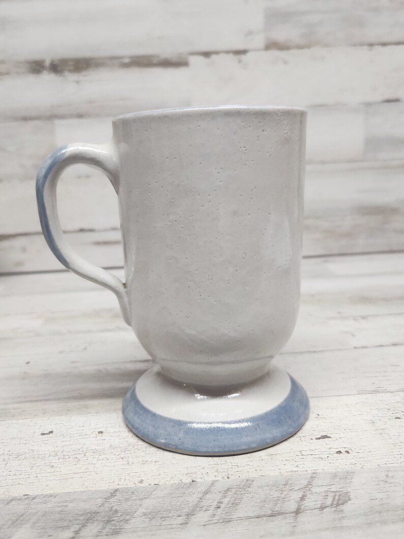 May include: A white ceramic mug with a blue band around the base. The mug has a handle and a rounded base.
