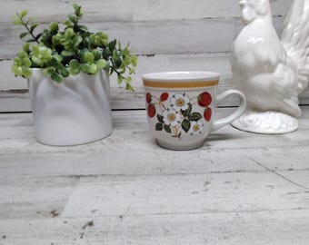 May include: A ceramic mug with a strawberry and floral design. The mug has a white base with a yellow band at the rim. The design features red strawberries and white flowers with green leaves. A white ceramic rooster and a white planter with greenery are also in the image.