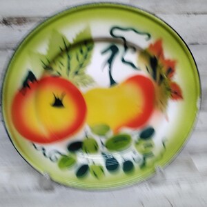 Peut inclure: Une assiette en &eacute;mail vintage avec un bord vert et un centre blanc. L'assiette pr&eacute;sente un motif peint de fruits, dont une pomme rouge et jaune et une poire jaune. Les fruits sont entour&eacute;s de feuilles vertes.