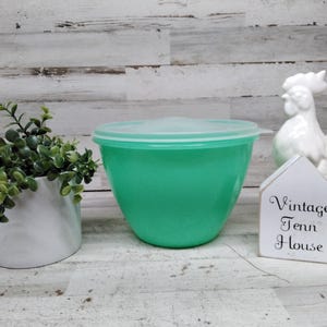 May include: A bright mint green plastic mixing bowl with a matching translucent lid. The bowl is positioned between a small white planter containing a green leafy plant and a white ceramic rooster figurine next to a small sign reading Vintage Tenn House.
