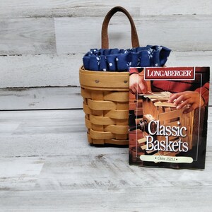 May include: A Longaberger Classic Baskets book with a brown woven basket with a blue and white fabric liner and a leather handle.