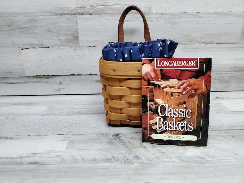 May include: A Longaberger Classic Baskets book with a brown woven basket with a blue and white fabric liner and a leather handle.