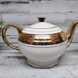 以下が含まれることがあります： 金色の取っ手、注ぎ口、蓋が付いた白と金のティーポット。 ティーポットの中央には金の帯があり、蓋には金のアクセントが付いています。 ティーポットはヴィンテージスタイルで、おそらくセラミック製です。
