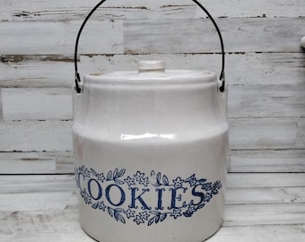 May include: A white ceramic cookie jar with a lid and a black metal handle. The jar features the word "COOKIES" in blue script surrounded by floral designs. The jar is on a light wood surface.