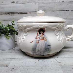 May include: A cream-colored ceramic chamber pot with a lid and a side handle. The exterior features embossed scrollwork and a central illustration of a person in a white dress with a red ribbon, accompanied by the text "Porky" and "Loomis".