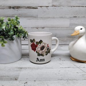 Puede incluir: Una taza de cerámica blanca con un diseño floral con flores rojas y blancas, y el nombre "Ann" en escritura negra. Junto a la taza hay una figura de pato de cerámica blanca y una maceta blanca con follaje verde.