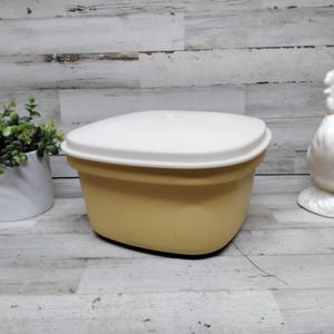 Puede incluir: Un recipiente de plástico amarillo mostaza vintage con tapa blanca. El recipiente es cuadrado con bordes redondeados. Un gallo de cerámica blanca y una pequeña planta en maceta están en el fondo.