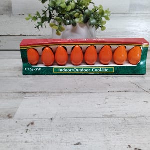 Puede incluir: Un paquete de ocho bombillas naranjas C7 ½-5W. Las bombillas tienen forma de huevo y están en una caja roja, verde y negra. La caja dice "Time At Home" e "Indoor/Outdoor Cool-lite". Una pequeña planta verde está en el fondo.