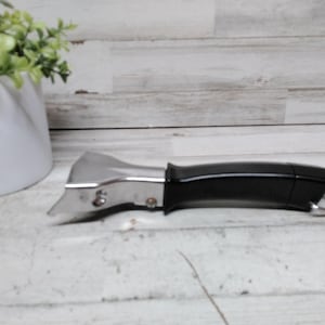 Può includere: Un apriscatole in acciaio inossidabile e nero con un anello argentato. L'apriscatole è appoggiato su una superficie di legno bianco. Un vaso di ceramica bianco con piante verdi è sullo sfondo.