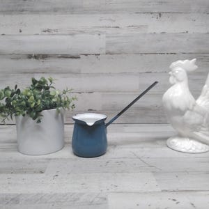 Könnte beinhalten: Eine weiße Keramik-Hahn-Statue, ein kleiner blau-weißer Emaille-Krug und ein weißer Keramik-Pflanztopf mit Grünzeug sind auf einer hellen Holzoberfläche angeordnet. Der Krug hat einen langen, dünnen Griff.