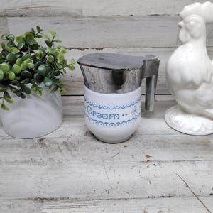 May include: A white ceramic creamer with a silver lid and handle, adorned with blue text that reads "Cream" and decorative blue accents. A white ceramic rooster and a small potted plant are also in the image.