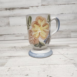 May include: A white ceramic mug with a blue rim and a floral design. The flowers are painted in shades of yellow, orange, and blue.