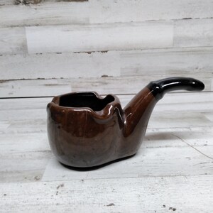 Könnte beinhalten: Eine braune Keramikpfeife in Form einer Tabakpfeife mit schwarzem Stiel. Die Pfeife ist als Aschenbecher konzipiert.