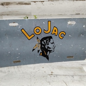 Könnte beinhalten: Rechteckiges, graues Metallkennzeichen mit dem Wort "LOJAC" in gelben Blockbuchstaben. Darunter ein schwarz-gelbes Profil eines amerikanischen Ureinwohners mit einer Feder. Die Platte hat vier Befestigungslöcher.