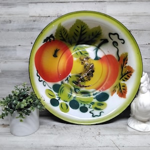 Op de afbeelding: Een vintage emaille kom met een witte binnenkant en een groene rand. De kom heeft een handgeschilderd ontwerp van twee oranje en gele pompoenen, groene bladeren en druiven. Rechts van de kom staat een klein wit keramieken hanenbeeldje.