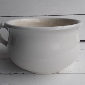 Pode incluir: Um penico de cerâmica branco com uma única pega. O penico é redondo e tem um acabamento liso e brilhante.