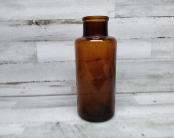 May include: A brown glass bottle with a wide mouth opening. The bottle is empty and has a slightly textured surface.