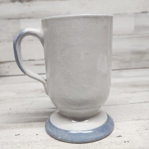 May include: A white ceramic mug with a blue band around the base. The mug has a handle and a rounded base.