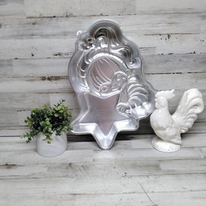 May include: A silver-colored, star-shaped cake pan in the shape of a woman's face and hair. A white ceramic rooster figurine and a small potted plant are also in the image.