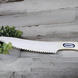 May include: A white plastic Kraft cheese knife with a serrated edge. The knife has a curved blade and a handle with a hole for hanging. The Kraft logo is printed in blue on the handle.