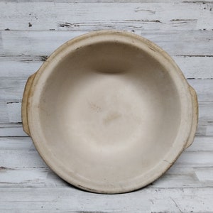 Könnte beinhalten: Eine beige, runde Keramikschüssel mit zwei seitlichen Griffen. Die Schüssel hat einen breiten Rand und ein glattes Inneres. Die Außenseite weist Gebrauchsspuren auf. Die Schüssel ist wahrscheinlich zum Kochen oder Servieren gedacht.