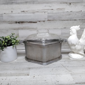 Puede incluir: Una fuente cuadrada plateada con tapa de cristal transparente, una figura de gallo de cerámica blanca y una maceta blanca con vegetación. La fuente tiene una base y asas decorativas.