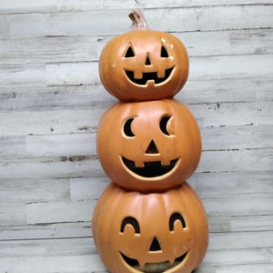 Può includere: Una pila di tre zucche arancioni a forma di jack-o'-lantern con facce intagliate. Le zucche sono impilate l'una sull'altra, con la zucca più grande in basso e la più piccola in alto.