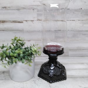 Op de afbeelding: Een heldere glazen hurricane kandelaar met een donkerrode basis en een zwarte decoratieve basis. Links van de kandelaar staat een kleine witte keramische pot met groene bladeren.