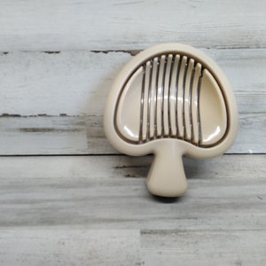 May include: A beige plastic egg slicer shaped like a mushroom with a handle and multiple metal blades.