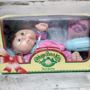 Może przedstawiać: Lalka Cabbage Patch Kids Newborn w żółtym pudełku z przezroczystym plastikowym okienkiem. Lalka ma na sobie różowy strój i zielone oczy. Pudełko zawiera butelkę i różowe opakowanie z logo Cabbage Patch Kids.