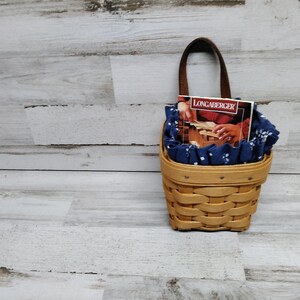 May include: A small, brown wicker basket with a blue and white fabric liner. The basket has a brown leather handle and a small book titled "Longaberger" inside.