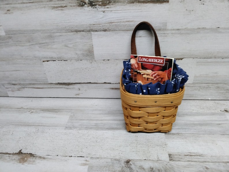 May include: A small, brown wicker basket with a blue and white fabric liner. The basket has a brown leather handle and a small book titled "Longaberger" inside.