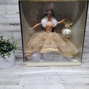 Może przedstawiać: Lalka Barbie w pudełku wystawowym. Lalka ma na sobie złotą suknię z białym futrzanym kołnierzem i trzyma ozdobę choinkową. Na pudełku widnieje napis "Celebration Forever".