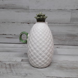 May include: White ceramic pineapple-shaped pitcher with a green handle and top. The pitcher has a textured surface and a decorative design. The handle and top are painted green, adding a pop of color to the white pitcher.