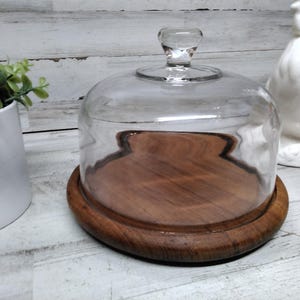 Puede incluir: Campana de cristal transparente con base de madera redondeada. La base de madera es de color marrón oscuro. Un pequeño asa de cristal en la parte superior. Un gallo de cerámica blanca en el fondo.