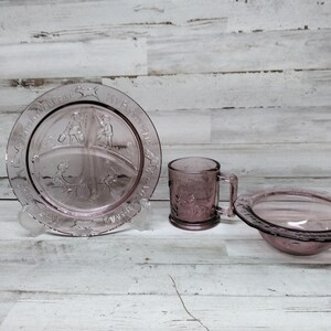 Puede incluir: Un juego de tres platos de vidrio rosa vintage, que incluye un plato dividido, una taza y un tazón. El plato tiene un diseño de niños jugando con un carro. La taza y el tazón tienen un diseño floral.