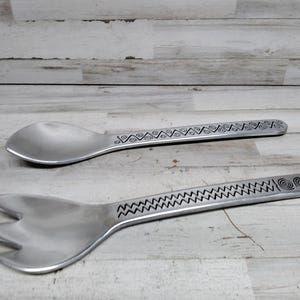 May include: Two silver serving utensils: a spoon and a fork. The spoon has a rounded bowl, and the fork has three tines. Both have etched handles with decorative patterns. The utensils are made of metal.