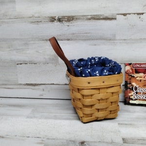 May include: A Longaberger Classic Baskets book with a woven basket with a blue and white fabric liner and a brown leather handle.